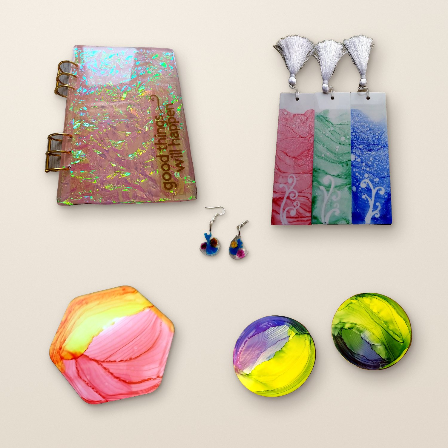 Hand-poured resin pieces laid out on a plaster surface: a holographic diary, alcohol ink bookmarks, teardrop earrings, a coral hexagon coaster and two round coasters