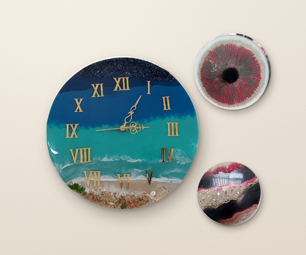 Three hand-poured resin wall pieces hung on a warm plaster wall: an ocean wall clock with gold numerals, the red Iris and the black and pink Black Magic geode